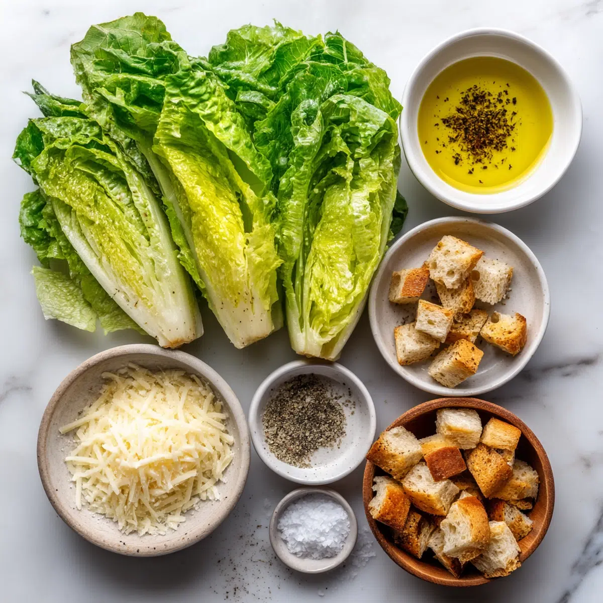 Caesar salad recipe ingredients laid out on a cutting board including romaine lettuce, parmesan cheese, bread cubes, olive oil, and garlic powder