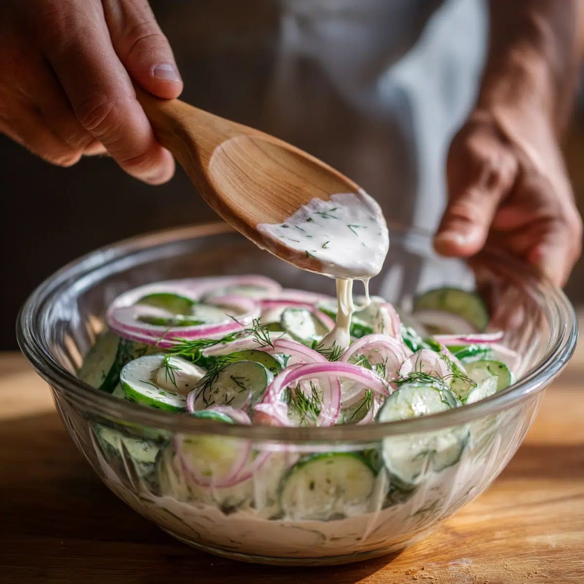 Creamy sour cream dressing being poured over sliced cucumbers and red onions in a glass mixing bowl